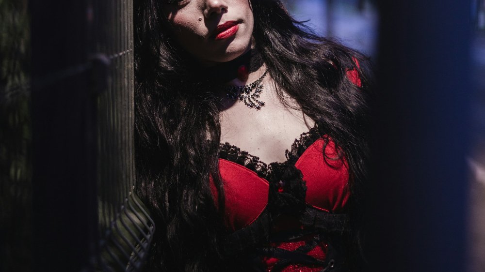 A pale woman dressed in a red and black corset with dark hair and red lipstick leaning against a stone column.